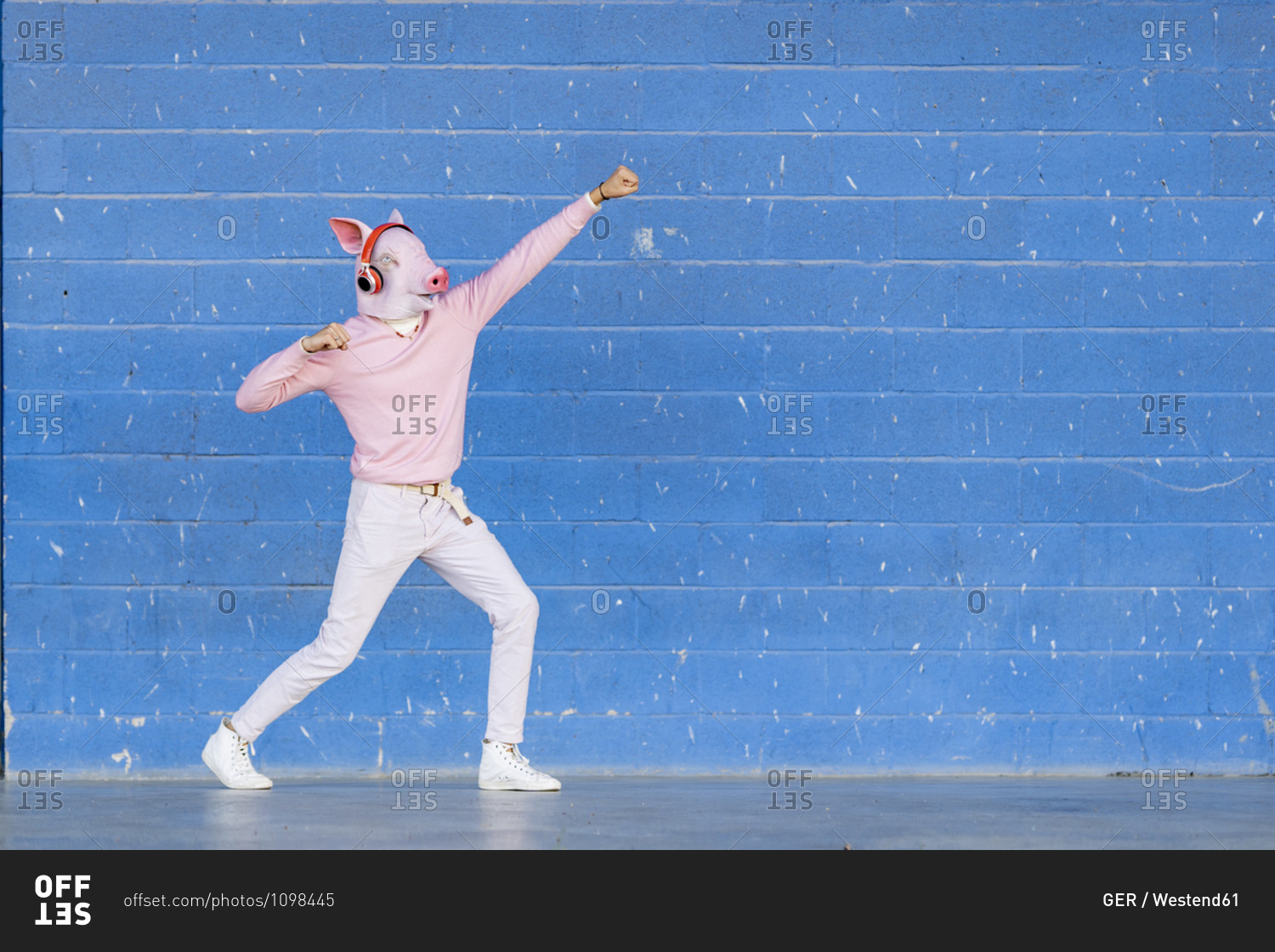 Fashionable young man wearing pig mask pretending to fly against blue wall