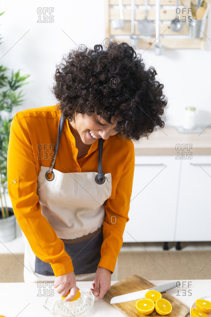 Smiling curly hair woman squeezing orange for making juice while standing in kitchen at home