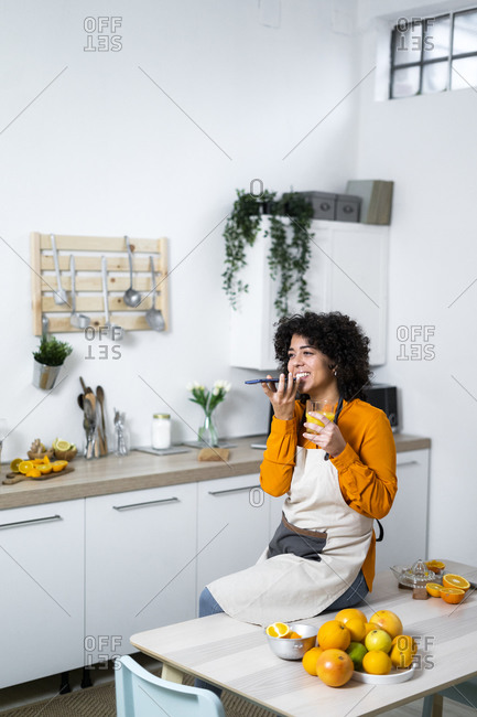 Smiling woman with orange juice talking on mobile phone while sitting on table by fruit at home