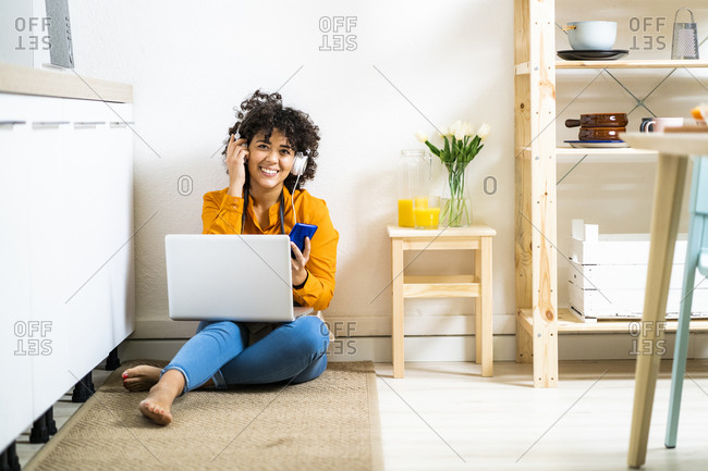 Young woman wearing headphones using mobile phone while sitting with laptop on floor at home