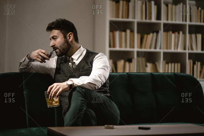 Portrait of bearded man relaxing on sofa with smoking pipe and glass of whiskey
