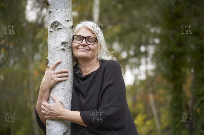 Smiling senior woman with eyes closed embracing tree trunk in public park