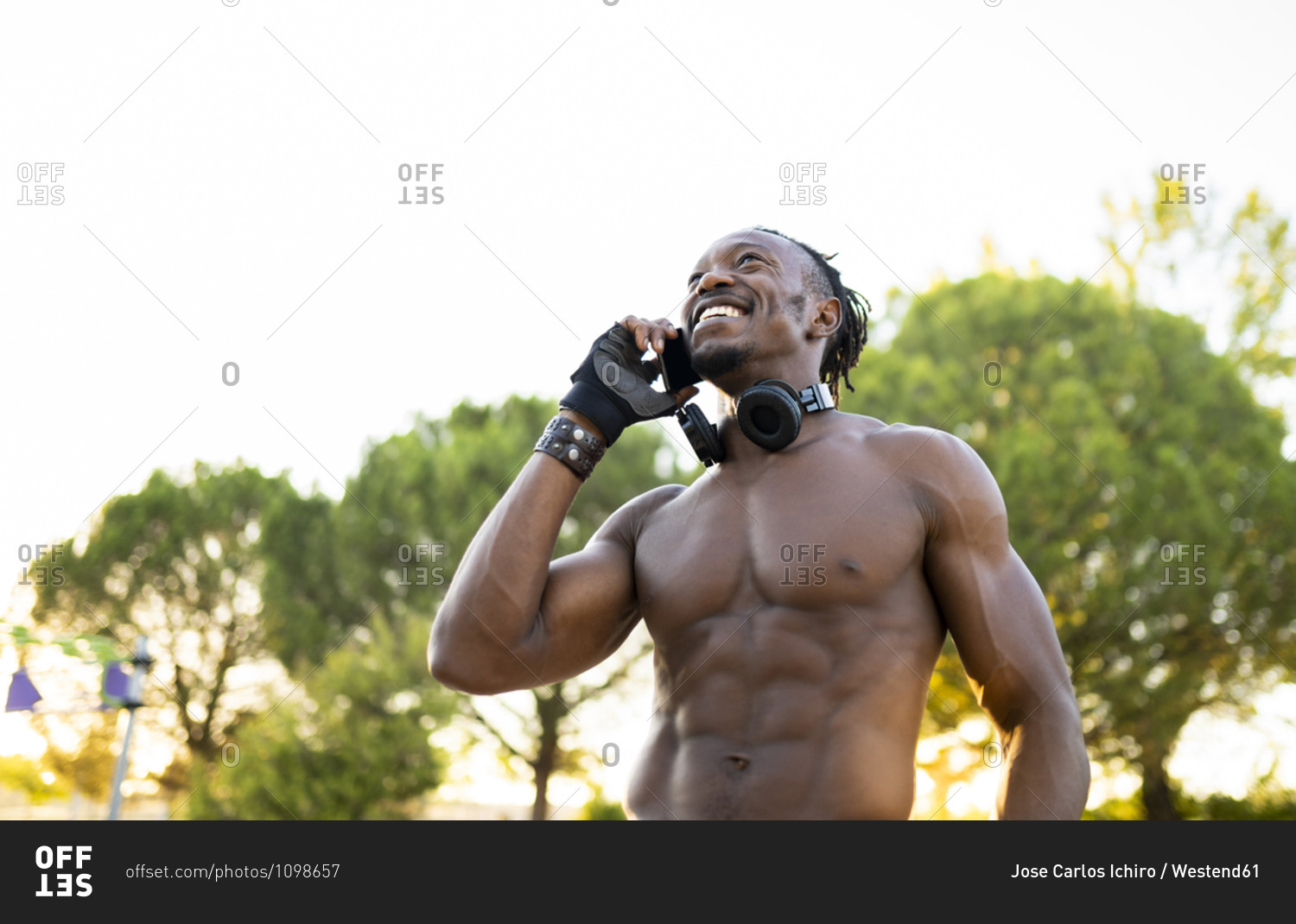 Shirtless sportsman with headphones talking o mobile phone while standing at park