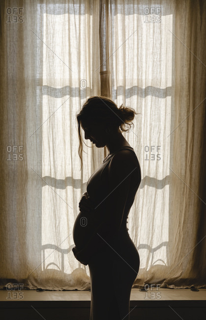 Pregnant woman touching belly while standing by window at home