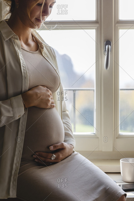 Pregnant woman with hands on stomach sitting by window at home