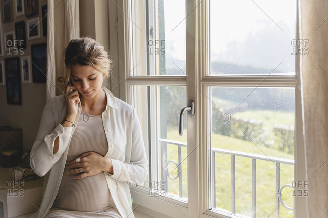 Young pregnant woman with hand on stomach talking through smart phone at home