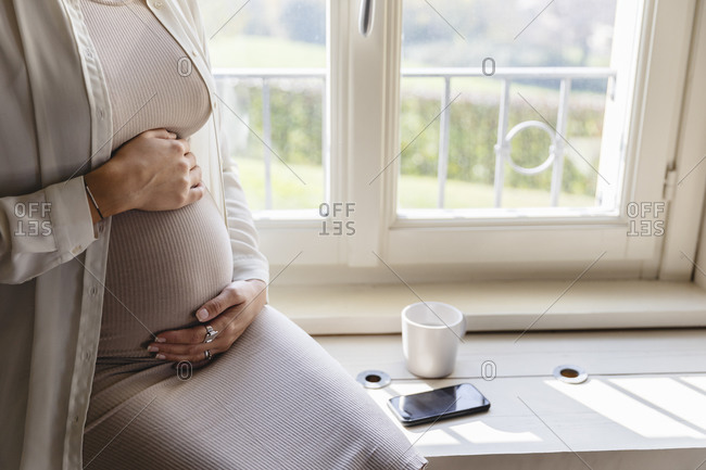 Pregnant woman hands on stomach sitting by window at home