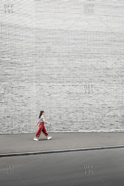 Woman wearing red pants walking on sidewalk by gray wall
