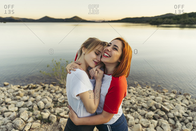 Happy woman embracing girlfriend while standing by lake during sunset