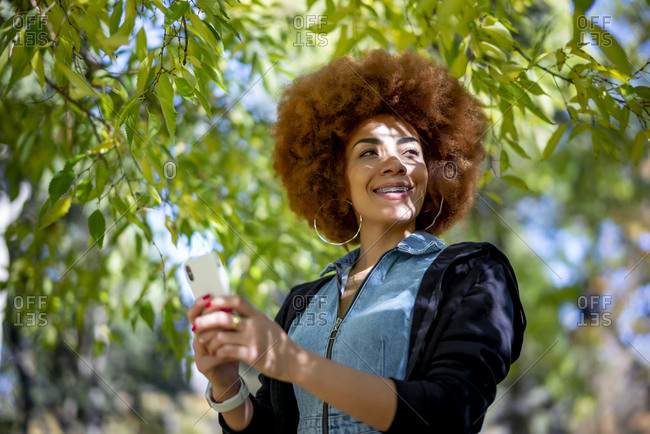 Smiling woman wearing hoop eating while holding mobile phone at public park