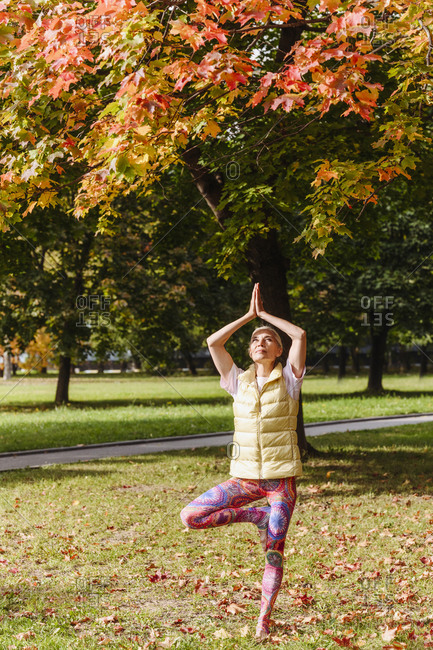 Mature woman balancing on one leg- practicing yoga in autumn park on sunny day