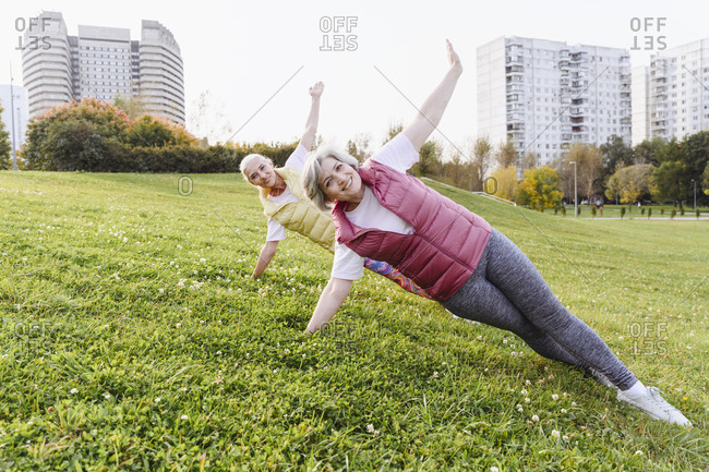 Smiling mature female friends exercising side plank on grass in public park