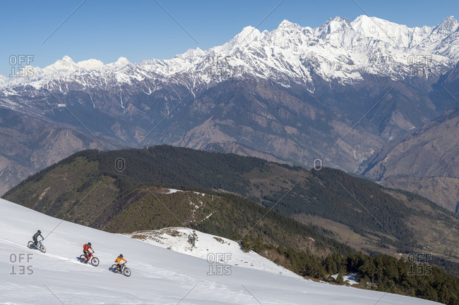 Mountain bikers descend a snow covered slope in the Himalayas with views of the Langtang range in the distance