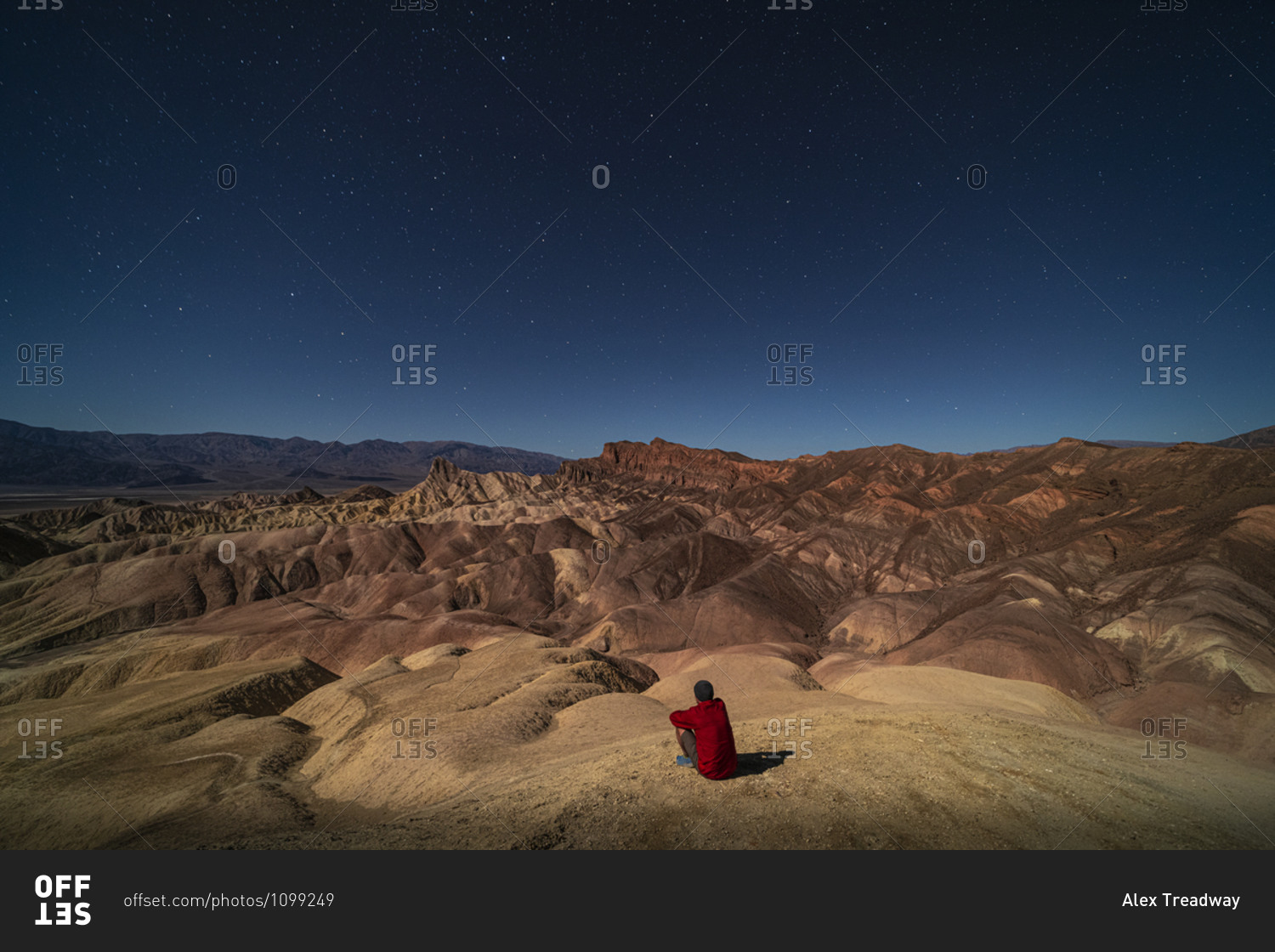 A man looking out at surreal rock formations caused by erosion at Zabriskie Point moonlit under a bright night sky full of stars