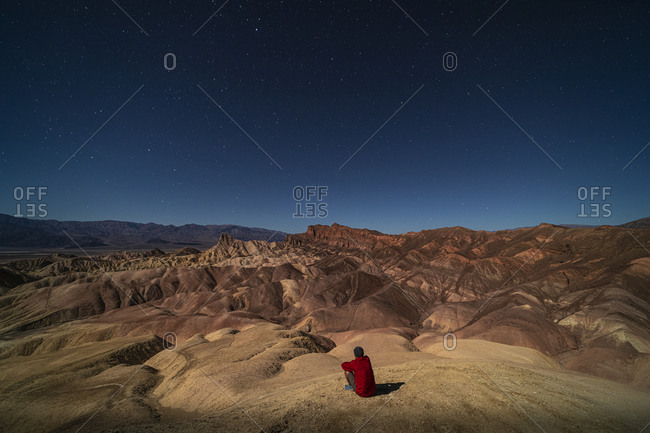 A man looking out at surreal rock formations caused by erosion at Zabriskie Point moonlit under a bright night sky full of stars