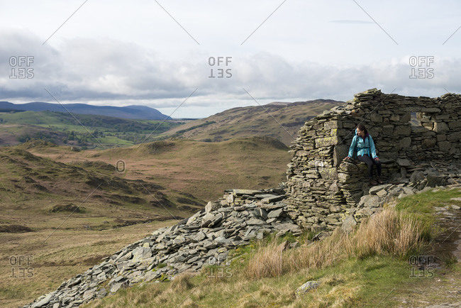 A woman rests on a crumbling stone house while climbing Place Fell in the English Lake District