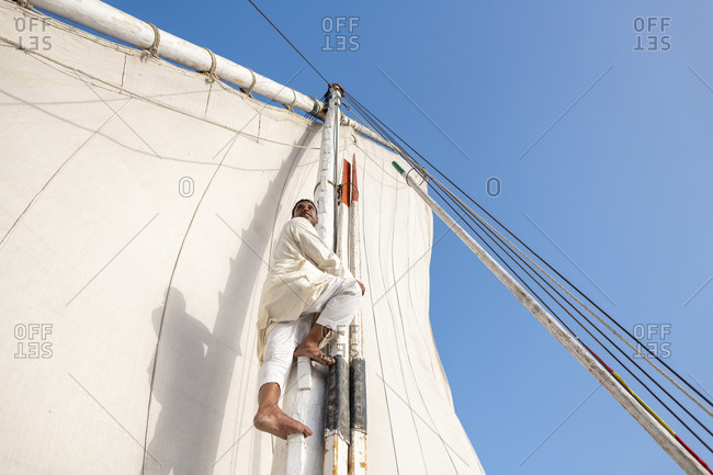 An Egyptian man stands on the bow of a traditional Felucca sailboat with wooden masts and cotton sails on the Nile river
