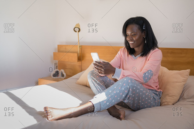 Happy black woman sitting in her bed wearing pajamas and looking at cell phone