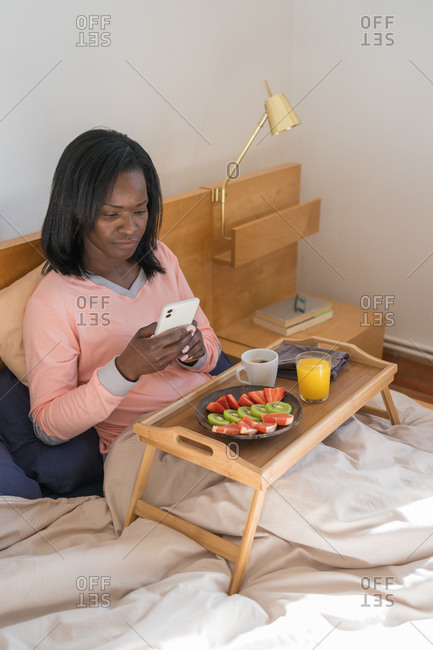 Black woman sitting in her bed wearing pajamas and looking at cell phone while eating a healthy breakfast