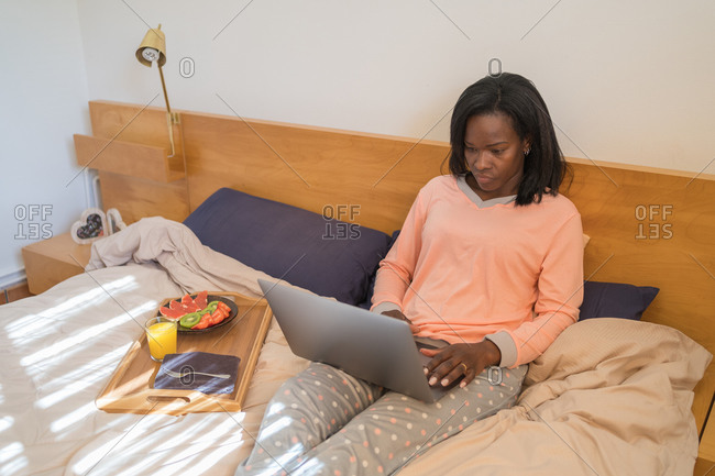 Black woman sitting in her bed wearing pajamas and using a laptop while eating a healthy breakfast