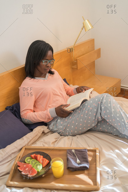 Black woman sitting in her bed wearing pajamas and reading a book while eating a healthy breakfast