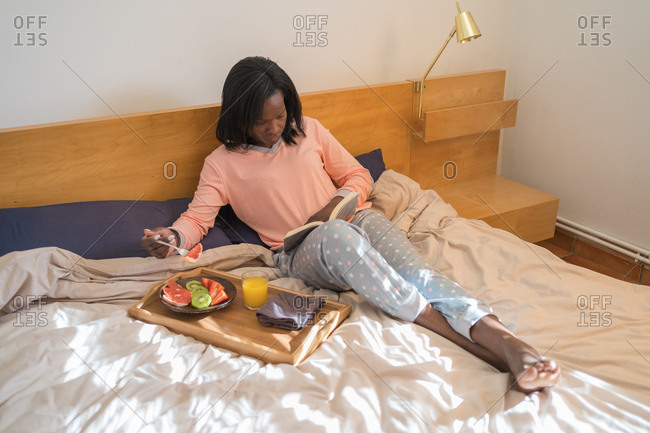 African American woman reading a book in her bed wearing pajamas and eating a healthy breakfast
