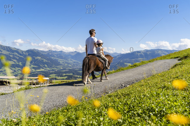 Father walking beside little daughter riding pony along alpine footpath