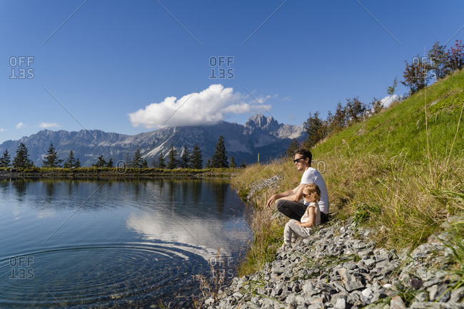 Father sitting with little daughter on shore of alpine lake