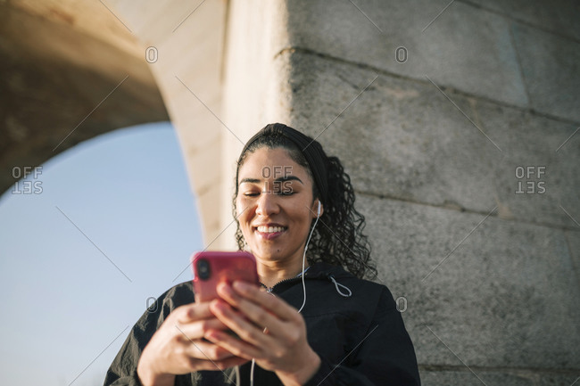 Smiling sportswoman using mobile phone while listening music against wall