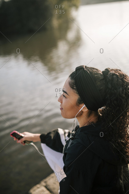 Female athlete with smart phone listening music while sitting by pond in public park