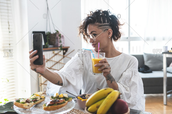 Portrait of young woman taking smart phone selfie while eating breakfast