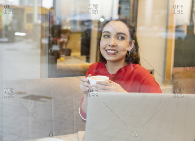 Smiling woman with coffee cup seen through glass in cafe