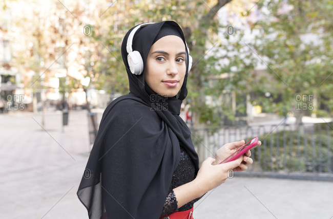 Portrait of young beautiful woman wearing hijab and headphones using smart phone