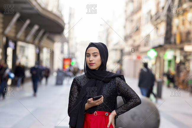 Portrait of young woman wearing black hijab standing on sidewalk with smart phone in hand