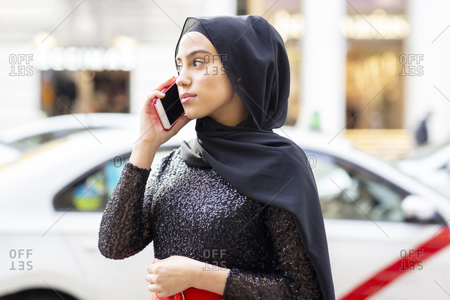 Portrait of young beautiful woman wearing black hijab talking on smart phone in middle of street