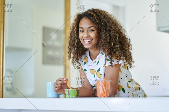 Smiling woman with guacamole sauce and carrots leaning on kitchen counter at home