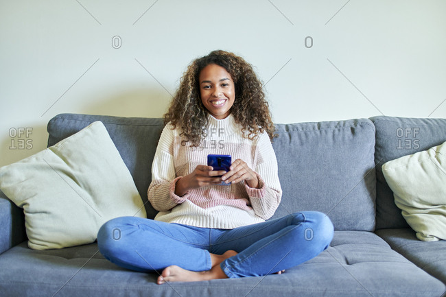Smiling afro young woman with mobile phone on sofa in living room at home