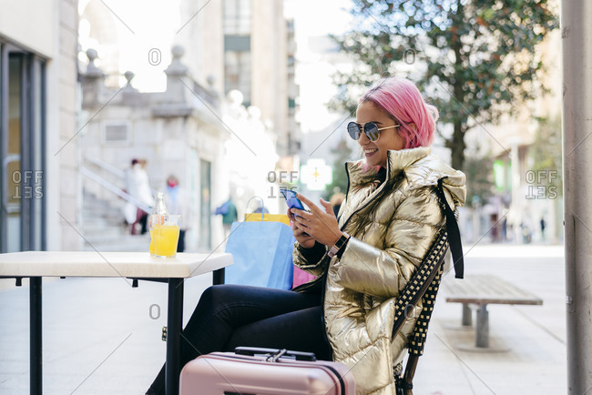 Smiling pink hair woman using mobile phone while sitting at sidewalk cafe