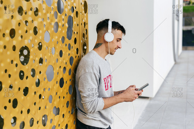 Young man wearing headphones using mobile phone while leaning on wall