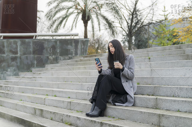 Female entrepreneur with reusable coffee cup using mobile phone on staircase