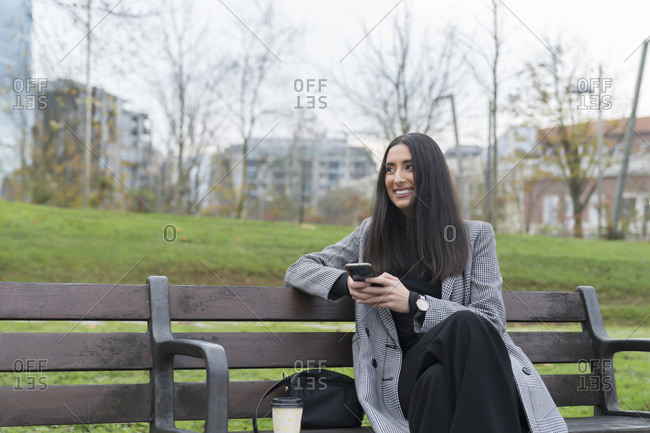 Smiling female professional with coffee cup looking away while holding mobile phone on park bench