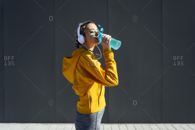 Female athlete wearing headphones drinking water while standing against gray wall