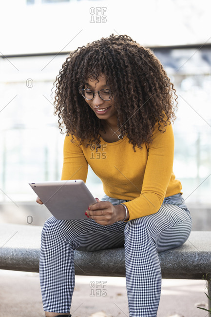 Smiling young woman with curly hair using digital tablet while sitting on bench