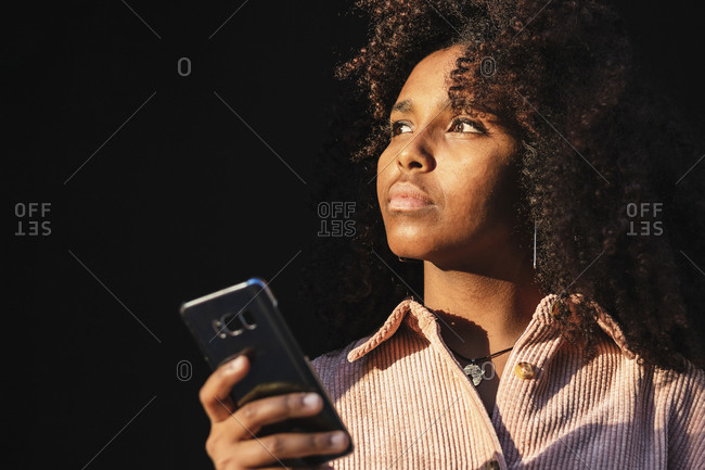 Thoughtful afro young woman holding mobile phone while looking away on sunny day