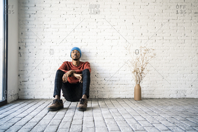Man wearing knit hat day dreaming while sitting by dried plat vase against white brick wall