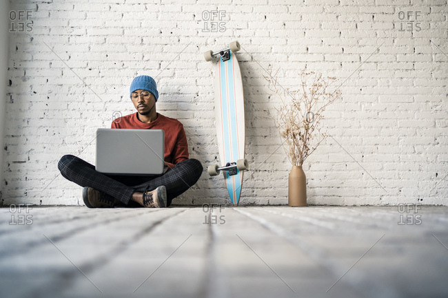 Businessman working on laptop while sitting by skateboard against white brick wall