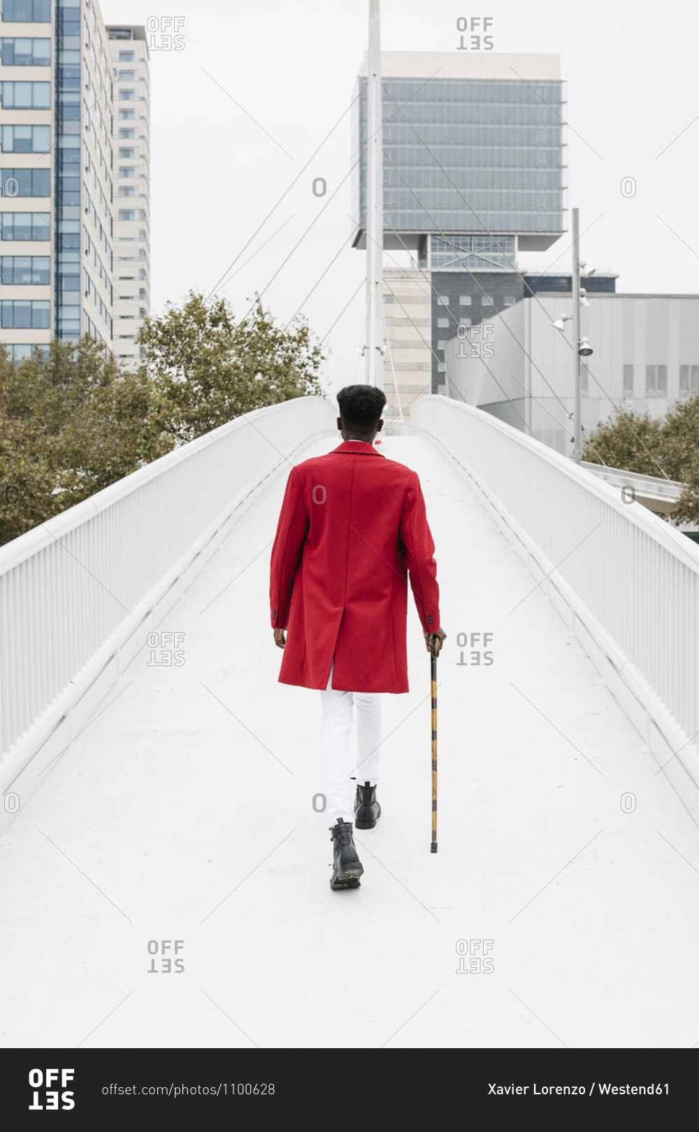 Man wearing red jacket walking with walking cane on bridge in city