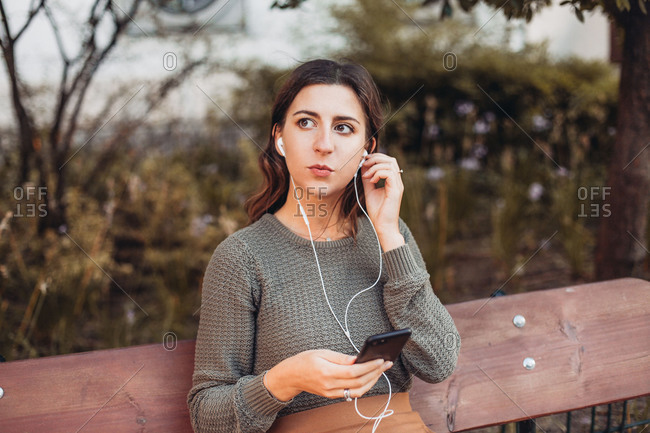 Young serious woman with earphones, a smartphone sitting on a bench
