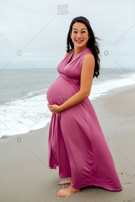 Very pregnant smiling Asian woman in pink dress at the beach