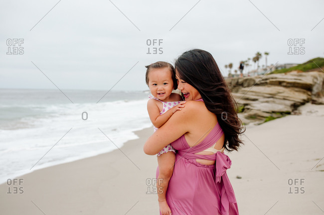 Smiling Asian mom holding happy year old daughter on beach near rocks